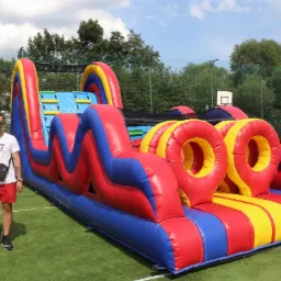 Kolorowy, dmuchany tor przeszkód z tunelem i obręczami na zielonej nawierzchni boiska, obok stoi mężczyzna w okularach słonecznych i czerwonych spodenkach.