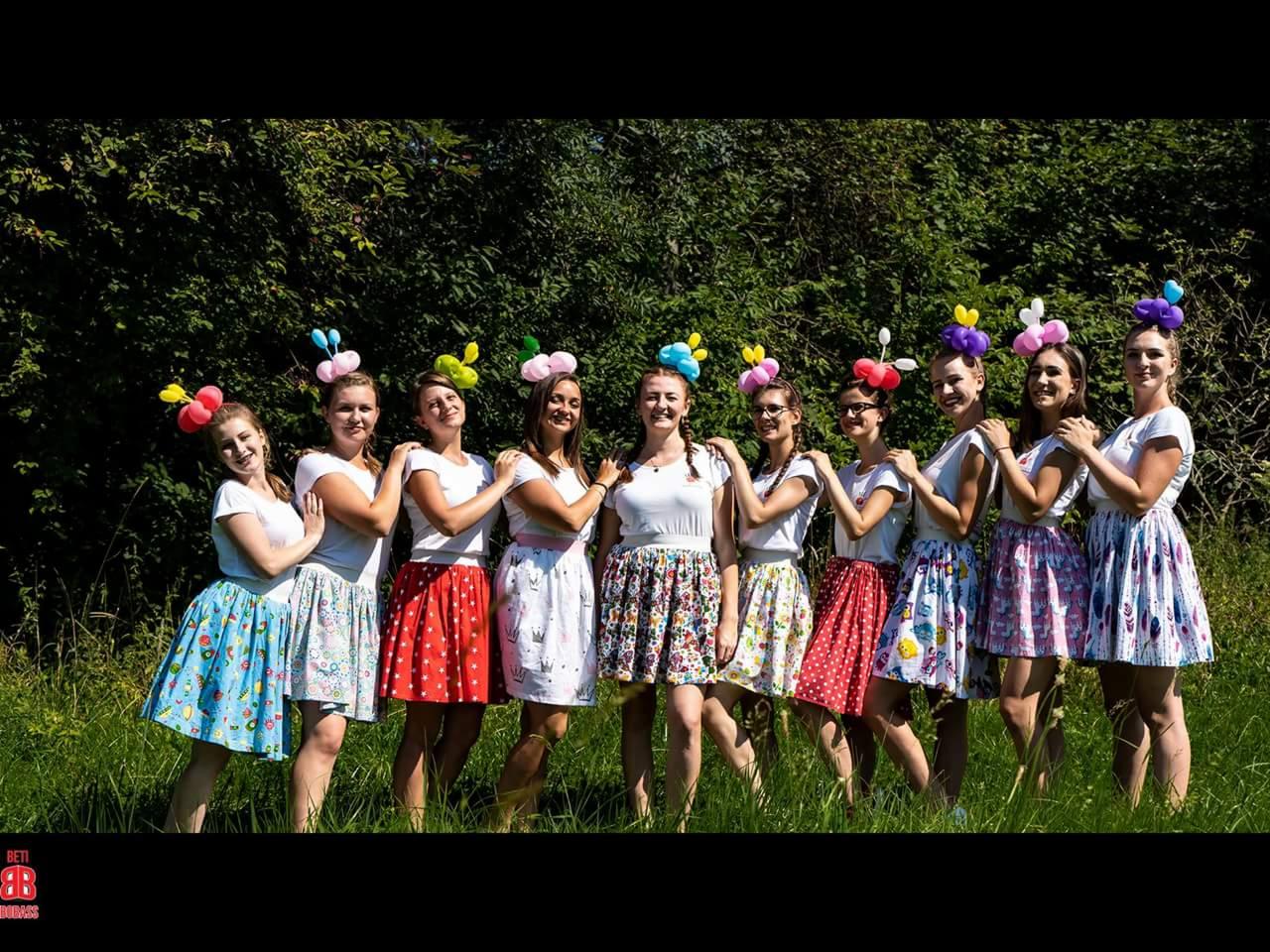 Grupa dziewcząt w kolorowych spódnicach i stroikach z balonów na głowach, pozujących na tle zieleni.