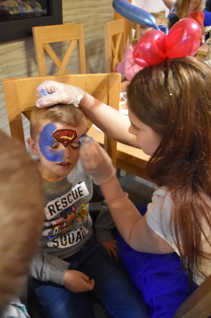 Malowanie twarzy chłopcu w motywie Supermana podczas imprezy urodzinowej, artystka w rękawiczkach aplikuje farbę na policzek dziecka.