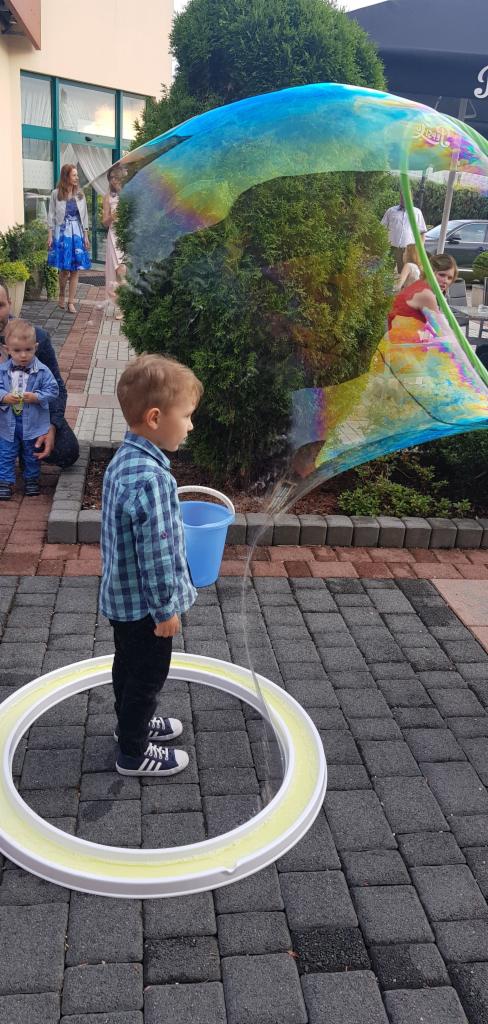 Chłopiec w niebieskiej koszuli w kratę stoi wewnątrz dużej, okrągłej obręczy do robienia baniek mydlanych, z których powstaje gigantyczna, tęczowa bańka.