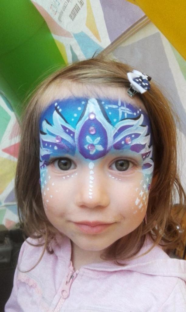 Dziewczynka z pomalowaną twarzą w niebiesko-fioletowe wzory z brokatem, przypominające koronę, z białą linią kropek na nosie i białymi wzorami na policzkach, w tle kolorowe dekoracje.