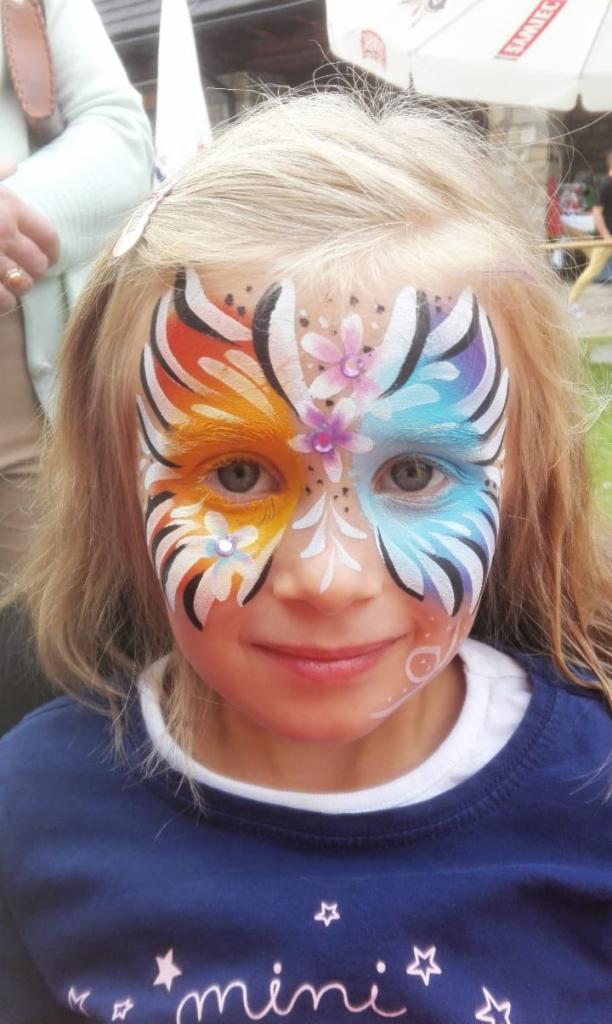 Dziewczynka z pomalowaną twarzą w motywy kwiatowe i geometryczne, z dominacją kolorów pomarańczowego i niebieskiego, na tle plenerowym.