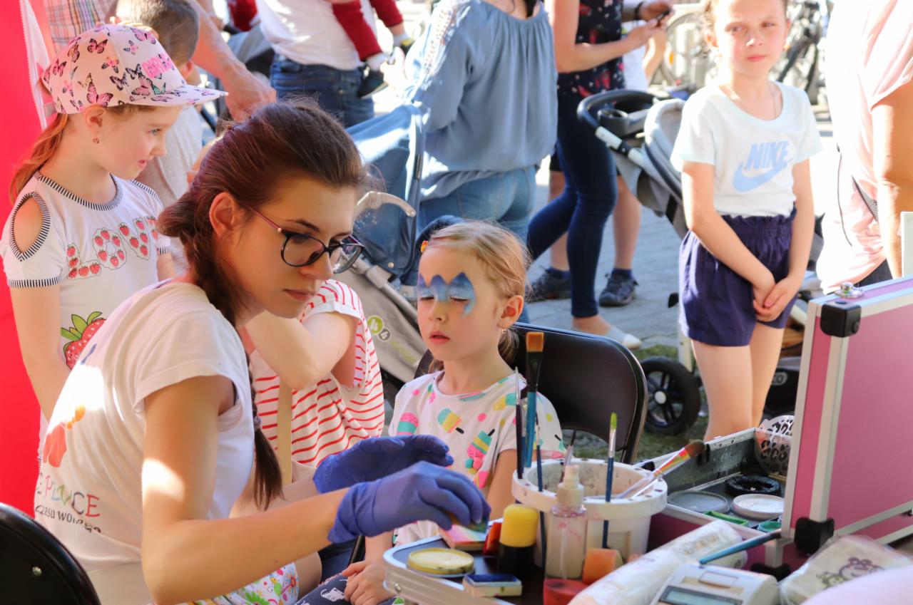 Młoda kobieta w niebieskich rękawiczkach maluje niebieskie wzory na twarzy dziewczynki siedzącej na krześle; na stole pędzle, farby i kosmetyczka; w tle inne dzieci i osoby.