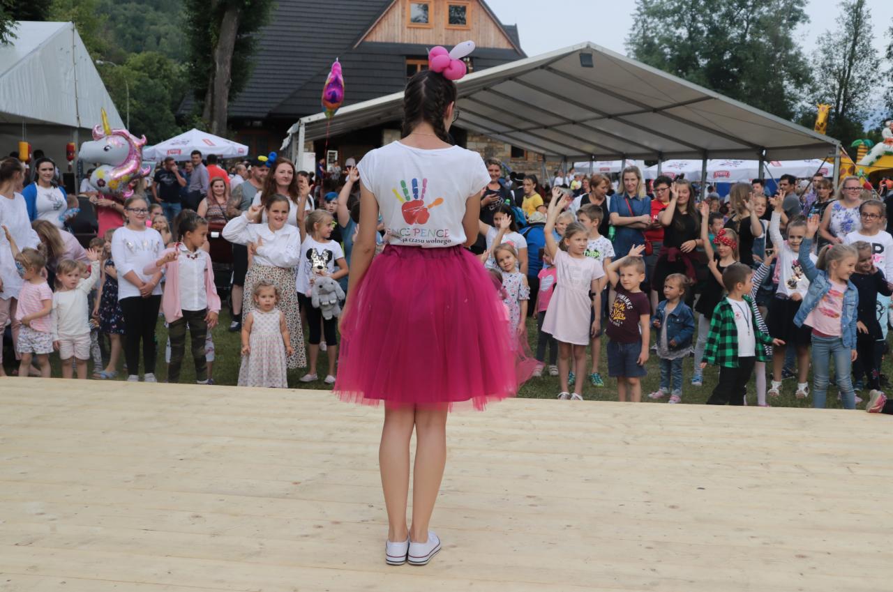 Animatorka w różowej spódnicy, tyłem do widza, na drewnianej scenie, przed grupą dzieci i dorosłych podczas imprezy plenerowej.