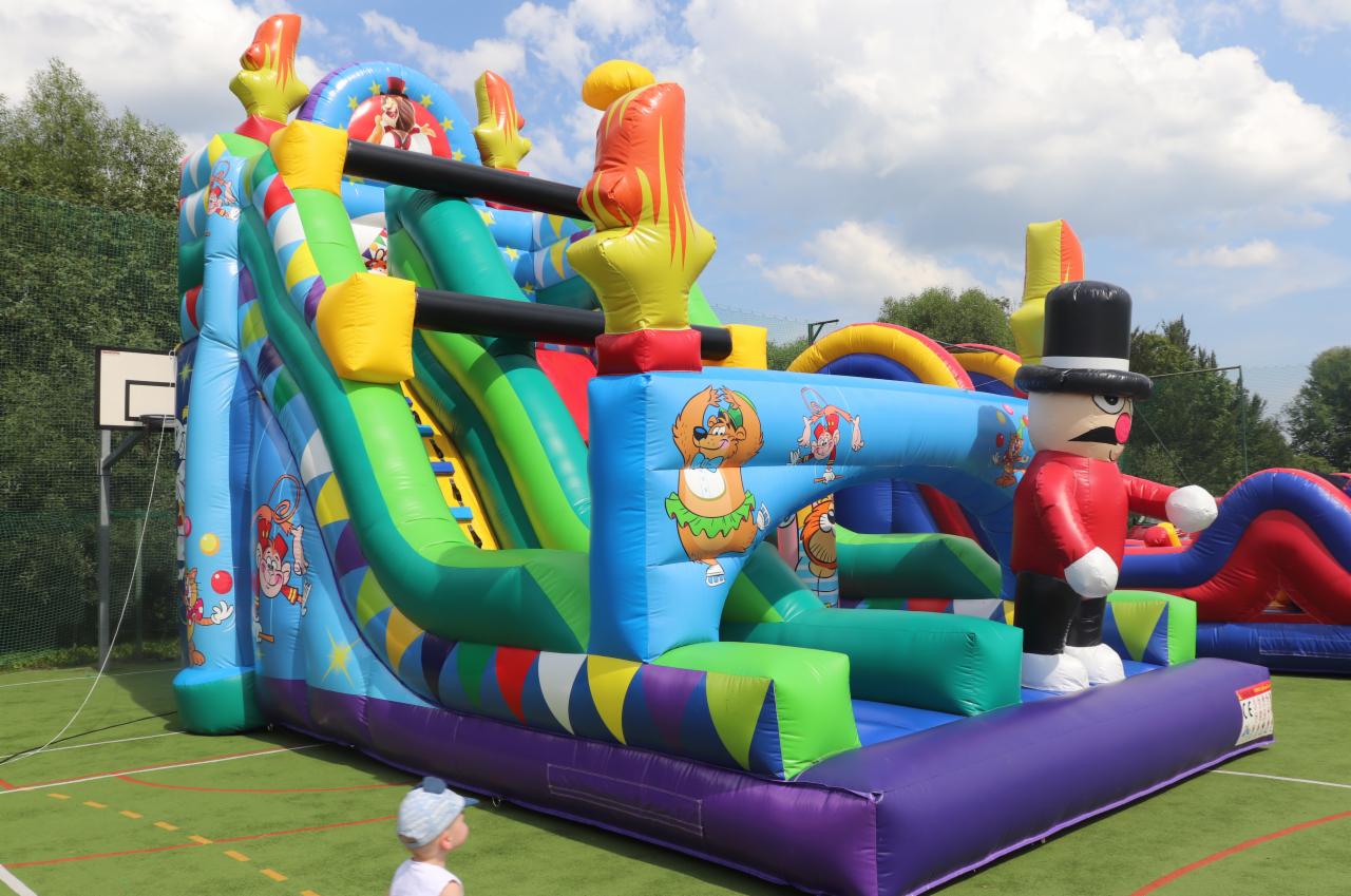 Kolorowa, nadmuchiwana zjeżdżalnia z motywami zwierzęcymi i postacią w cylindrze, ustawiona na zielonym boisku. W tle widoczne drzewa i błękitne niebo z chmurami.