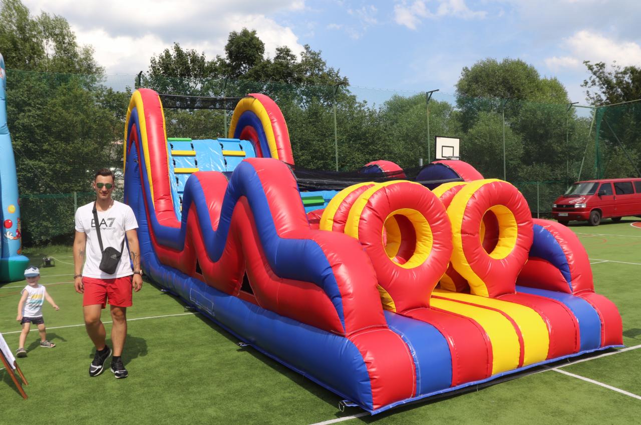 Kolorowy, dmuchany tor przeszkód z tunelem i obręczami na zielonej nawierzchni boiska, obok stoi mężczyzna w okularach słonecznych i czerwonych spodenkach.