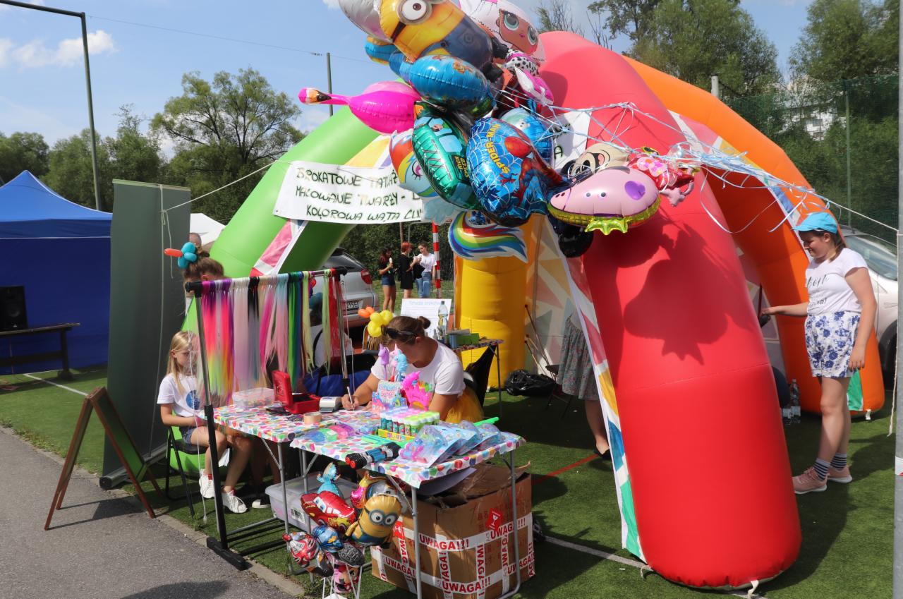 Stoisko z atrakcjami dla dzieci na świeżym powietrzu: kolorowe balony, dmuchana brama, stanowisko do malowania twarzy i zaplatania kolorowych pasemek, dziewczynka siedząca przy stoliku.