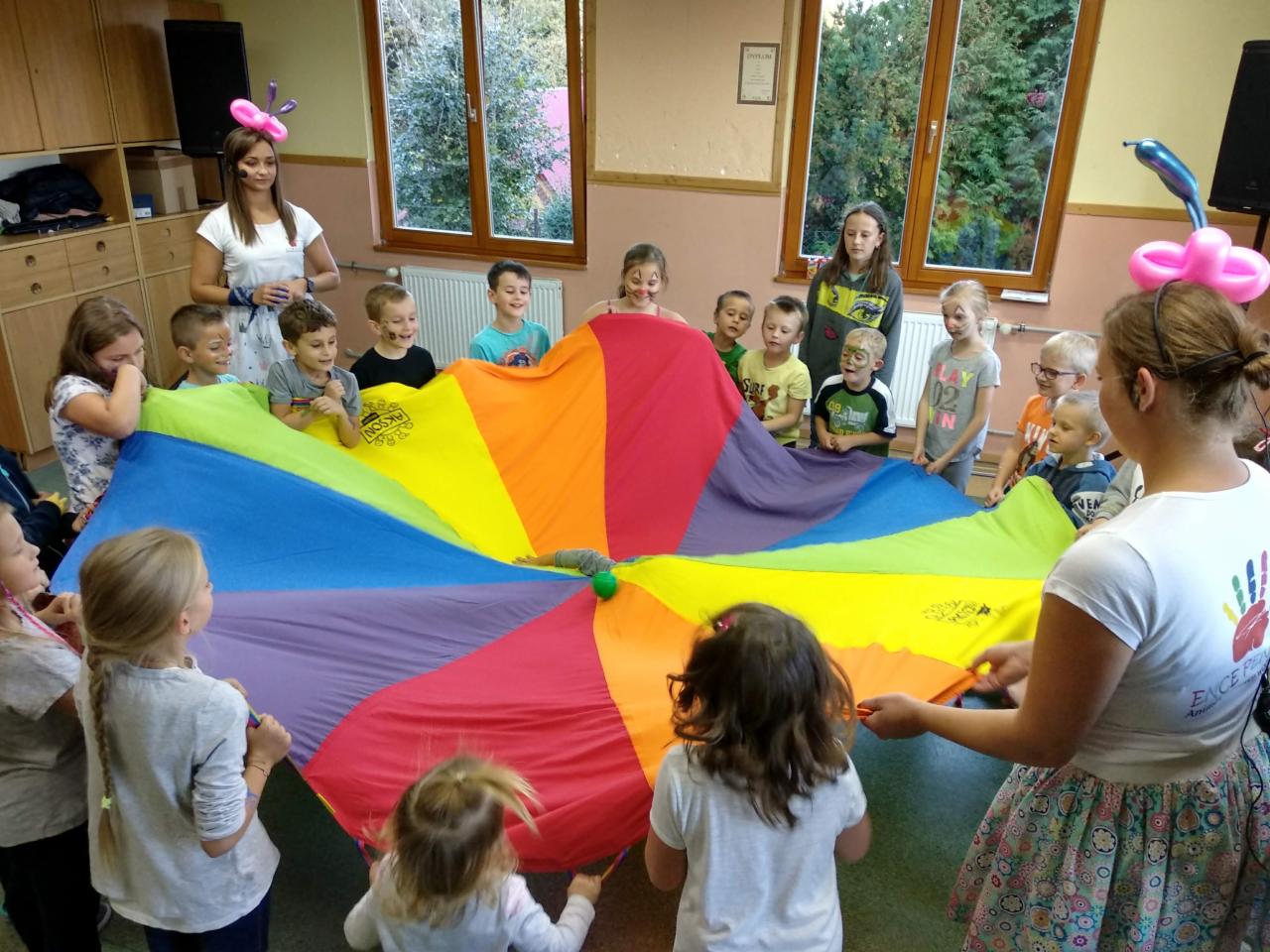 Grupa dzieci z pomalowanymi twarzami bawi się kolorową chustą animacyjną w sali z oknami i szafkami, nadzorowana przez dwie animatorki.