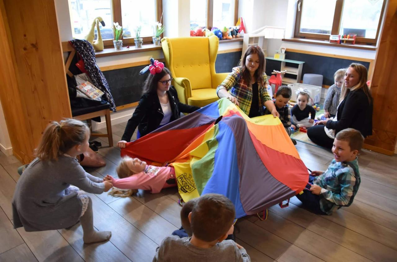 Grupa dzieci i dwie animatorki bawią się kolorową chustą animacyjną w jasnym pomieszczeniu z drewnianą podłogą; jedno dziecko leży pod chustą, pozostałe siedzą wokół.