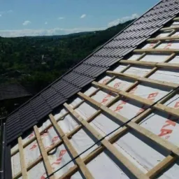 Częściowo pokryty dach z ciemnej dachówki, widoczna konstrukcja drewniana i membrana dachowa, w tle zalesione wzgórza.