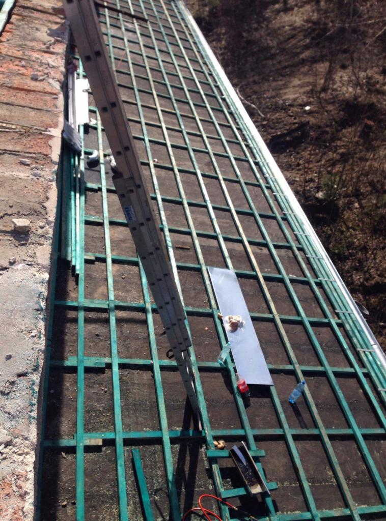 Konstrukcja dachu z zielonymi łatami, drabina oparta o ścianę, blacha, butelki i narzędzia na dachu podczas prac.