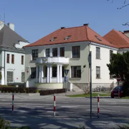 Elewacja dwupiętrowego budynku z balkonem wspartym na kolumnach, widoczny szyld 'RAD', obok zaparkowany czerwony samochód i rowery.