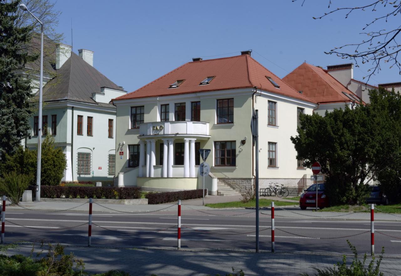 Elewacja dwupiętrowego budynku z balkonem wspartym na kolumnach, widoczny szyld 'RAD', obok zaparkowany czerwony samochód i rowery.