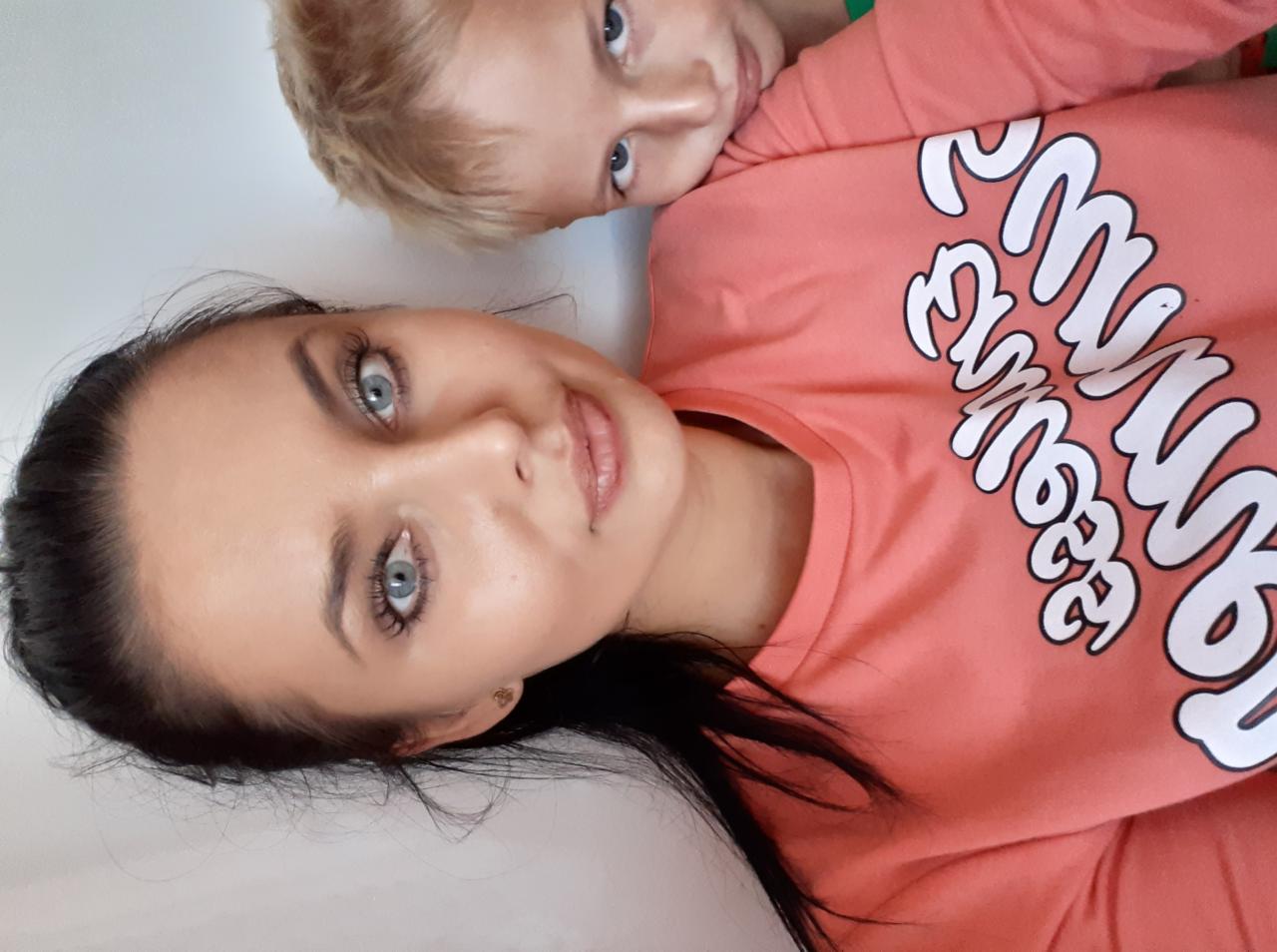 Uśmiechnięta kobieta z ciemnymi włosami i jasnymi oczami w koralowej bluzce z chłopcem o blond włosach, leżącym za nią. Selfie z bliska.