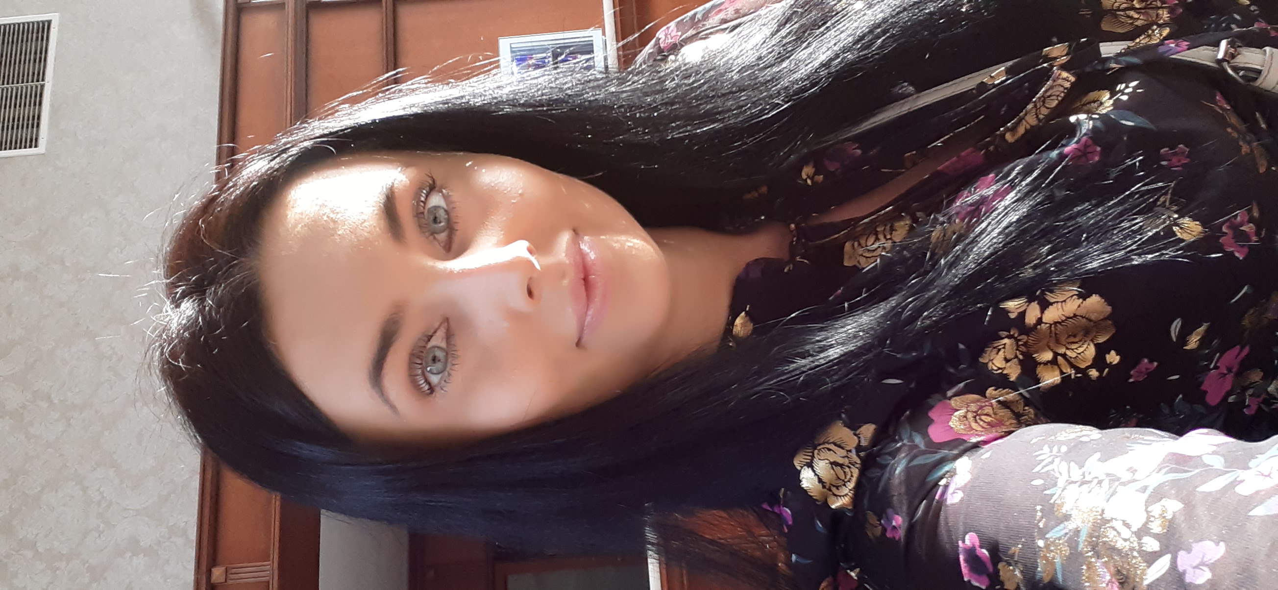 Portret kobiety o jasnych oczach, z długimi, czarnymi włosami i w kwiecistej bluzce, ustawiony pod kątem na tle jasnej ściany.