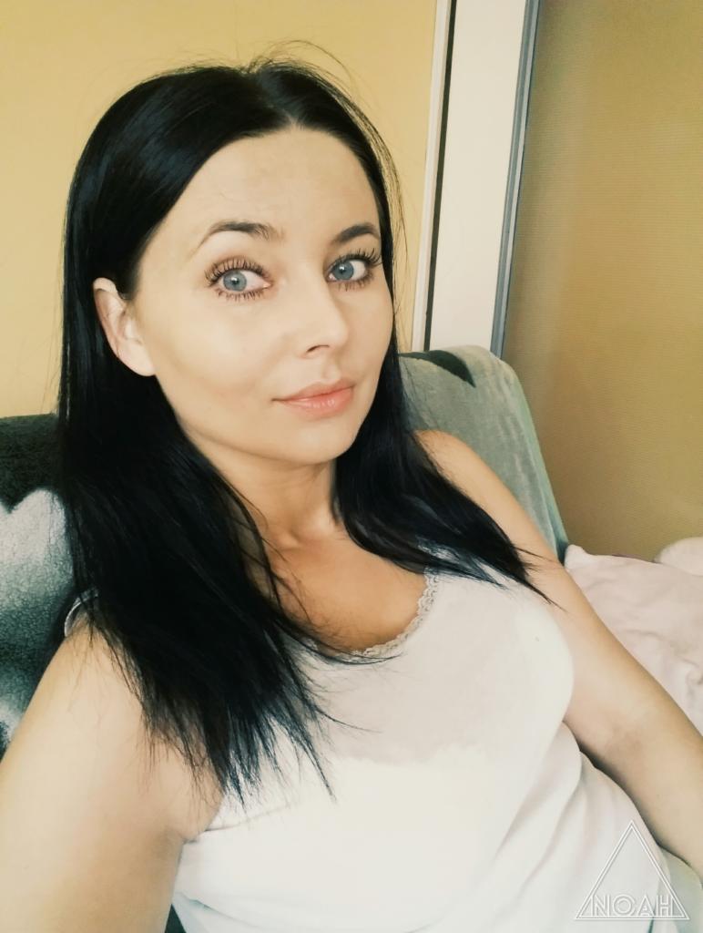 Portret kobiety z ciemnymi włosami i jasnymi oczami, ubrana w biały podkoszulek na tle pokoju.