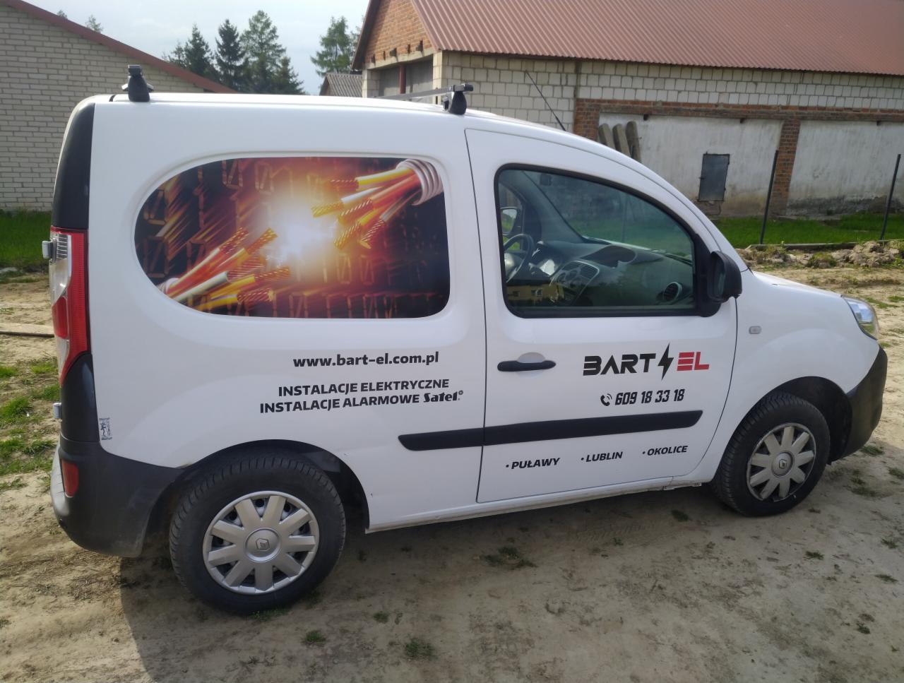 Biały samochód dostawczy z grafiką przedstawiającą wiązkę kabli elektrycznych i logo firmy BART-EL oferującej instalacje elektryczne i alarmowe, zaparkowany na piaszczystym podłożu przed budynkiem.