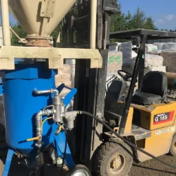 Niebieskie urządzenie do obróbki strumieniowo-ściernej ustawione obok żółtego wózka widłowego na zewnątrz, w tle worki z materiałami budowlanymi.