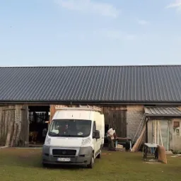 Nowy, grafitowy dach z blachy trapezowej na ceglanym budynku gospodarczym, z widocznym samochodem dostawczym i kurami na podwórku.