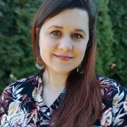 Portret uśmiechniętej brunetki z zielonymi oczami, w bluzce w kwiaty, z ozdobnymi kolczykami na tle zielonych krzewów.