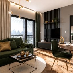 AUMÜLLERDESIGN Studio Projektowe - Elegancki salon z ciemnozieloną sofą narożną, złotymi poduszkami i wazonem, drewnianą podłogą w jodełkę. Duże okno z widokiem na miasto, czarna panelowa ściana RTV i okrągły stół.