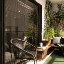 AUMÜLLERDESIGN Studio Projektowe - Elegancki balkon z drewnianą ławką, zielonymi poduszkami i bujną roślinnością w podświetlanych donicach, z widokiem na jadalnię z parkietem w jodełkę i marmurowym blatem.