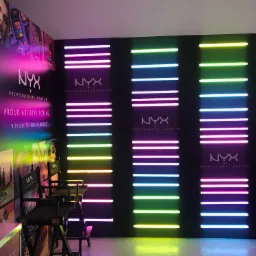 Stanowisko do makijażu marki NYX z neonowymi lampami w kolorach różowym, żółtym i zielonym, czarne krzesła reżyserskie z logo, ekspozycja kosmetyków na ścianie.