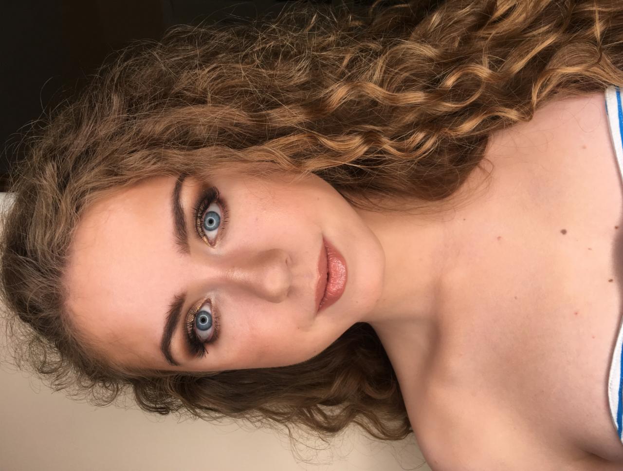 Portret młodej kobiety z profesjonalnym makijażem oczu w odcieniach złota i brązu, podkreślającym jej niebieskie oczy oraz z błyszczącymi ustami, z kręconymi włosami.