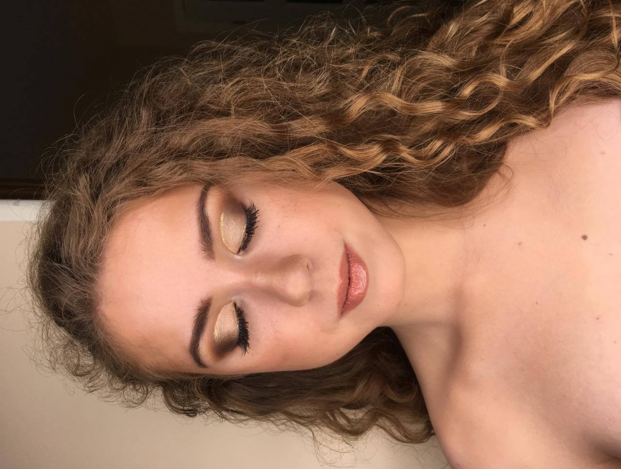 Portret kobiety z zamkniętymi oczami, prezentujący złoty, błyszczący cień do powiek i delikatny błyszczyk na ustach, podkreślone kręcone włosy.