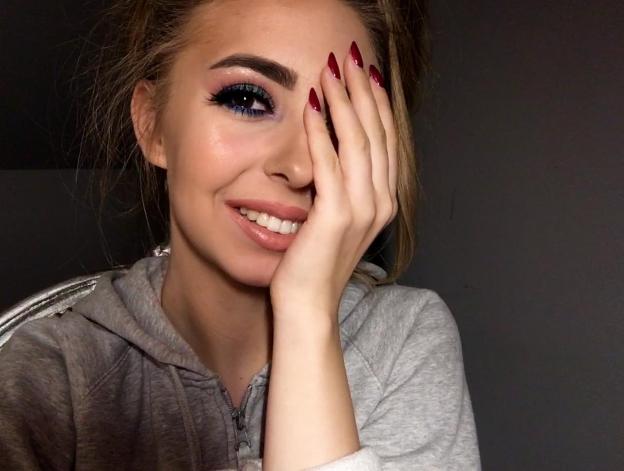 Uśmiechnięta młoda kobieta z intensywnym, niebiesko-różowym makijażem oczu i długimi, ciemnoczerwonymi paznokciami, zakrywająca część twarzy dłonią.