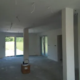 Wnętrze w trakcie prac wykończeniowych: świeżo pomalowane ściany i filary w jasnym odcieniu, widoczne wystające przewody elektryczne z sufitu, karton z farbą na podłodze w surowym stanie.