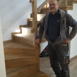 Stolarz prezentuje drewniane schody spiralne z oświetleniem punktowym, widoczny worek narzędziowy i narzędzia w tle.