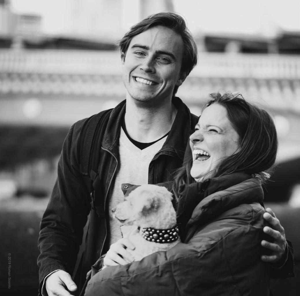 Czarno-białe zdjęcie uśmiechniętej pary z psem w obroży z ćwiekami, kobieta trzyma psa, oboje śmieją się, mężczyzna obejmuje kobietę.