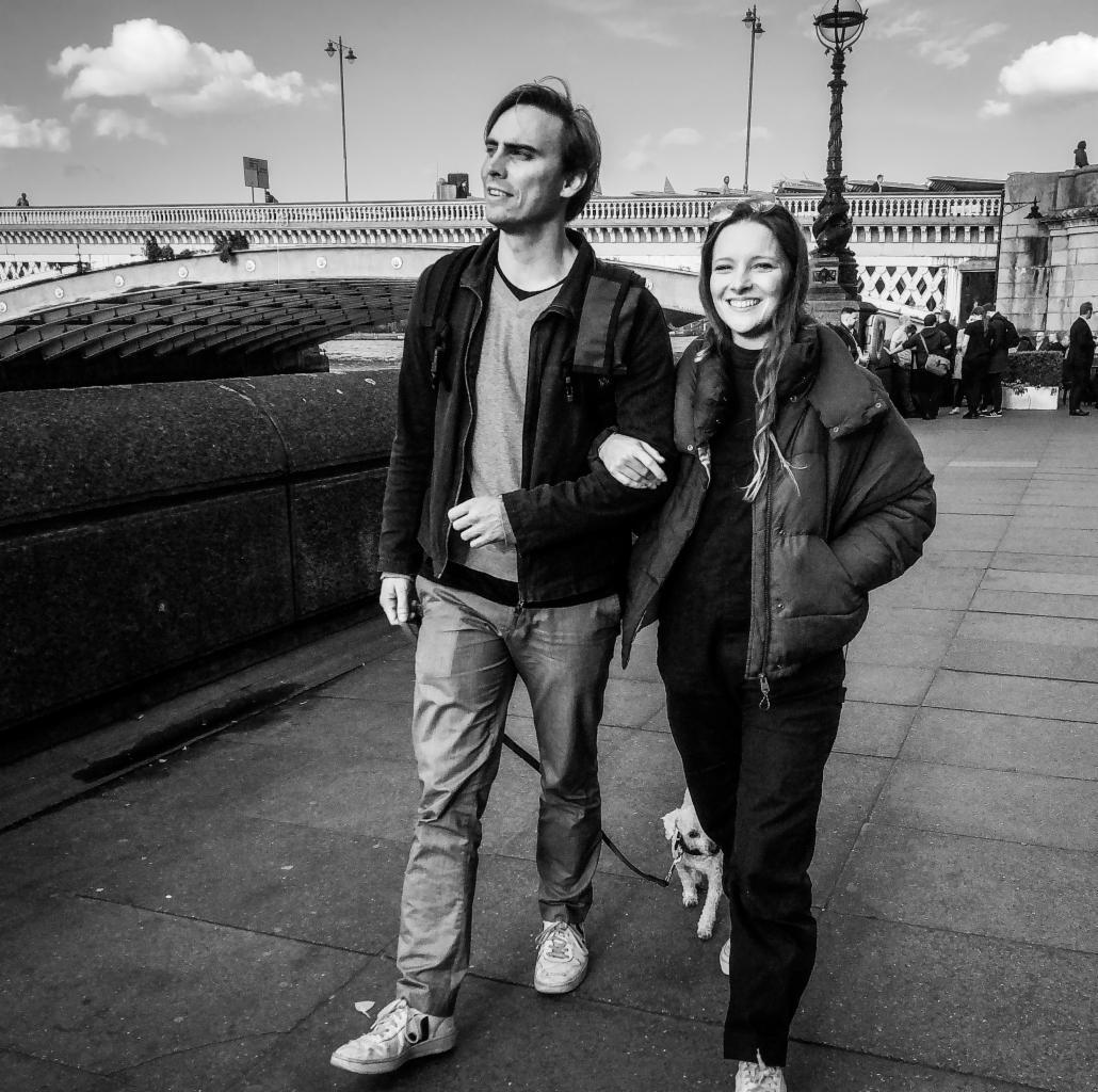 Czarno-białe zdjęcie pary z psem spacerującej po chodniku w London, z mostem i latarniami w tle.