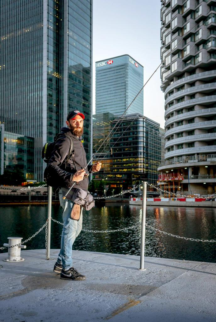 Mężczyzna z brodą w czapce i kurtce, z plecakiem i wędką, stoi na betonowym nabrzeżu w otoczeniu nowoczesnych wieżowców, wędkuje w miejskim krajobrazie.