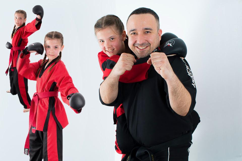 Uśmiechnięty mężczyzna w czarnym kimonie z czarnym pasem, dźwigający na plecach uśmiechniętą dziewczynkę w czerwonym kimonie, obok dziewczynka w czerwonym kimonie w postawie gotowości do walki,...