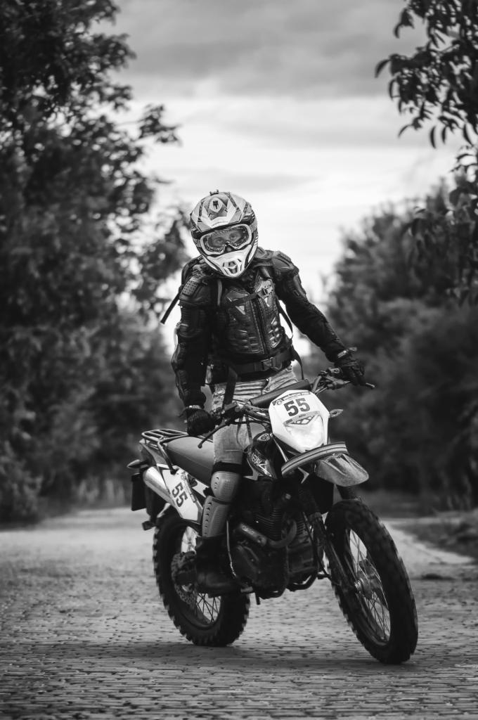 Czarno-białe zdjęcie motocyklisty w pełnym stroju ochronnym, stojącego na motocyklu crossowym z numerem 55 na tle leśnej ścieżki.