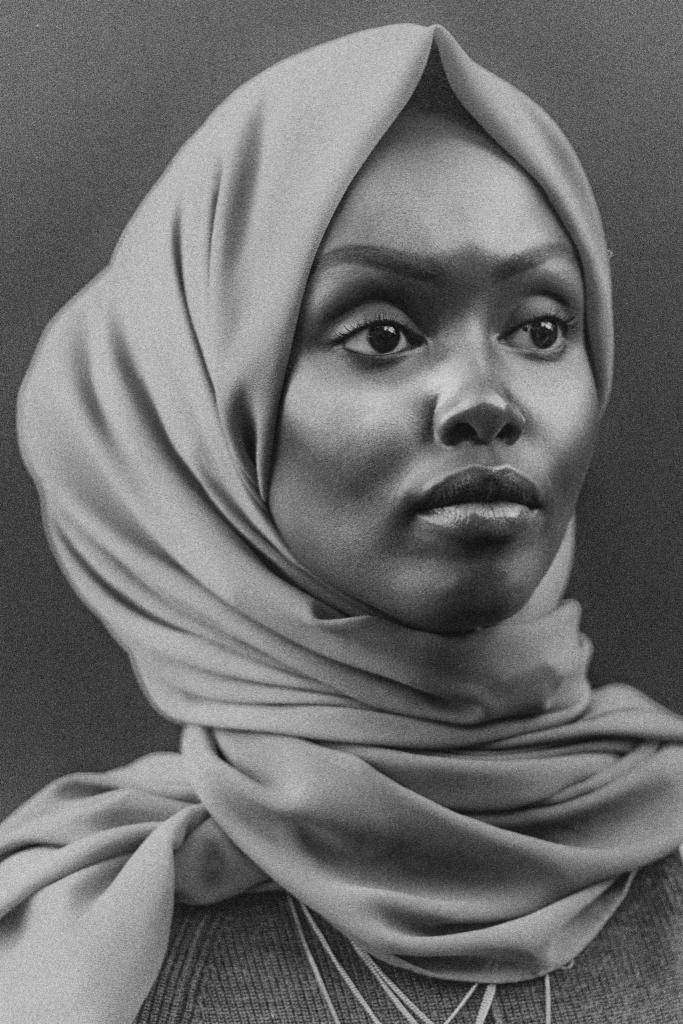 Czarno-biały portret kobiety z ciemną karnacją, ubranej w delikatny, drapowany szal zakrywający włosy, z wyrazem skupienia w oczach i subtelnym blaskiem na ustach.