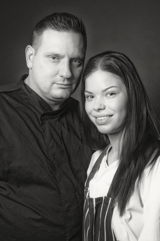 Portret biznesowy w czerni i bieli: mężczyzna w czarnej koszuli i kobieta w pasiastym fartuchu, oboje patrzą w obiektyw.