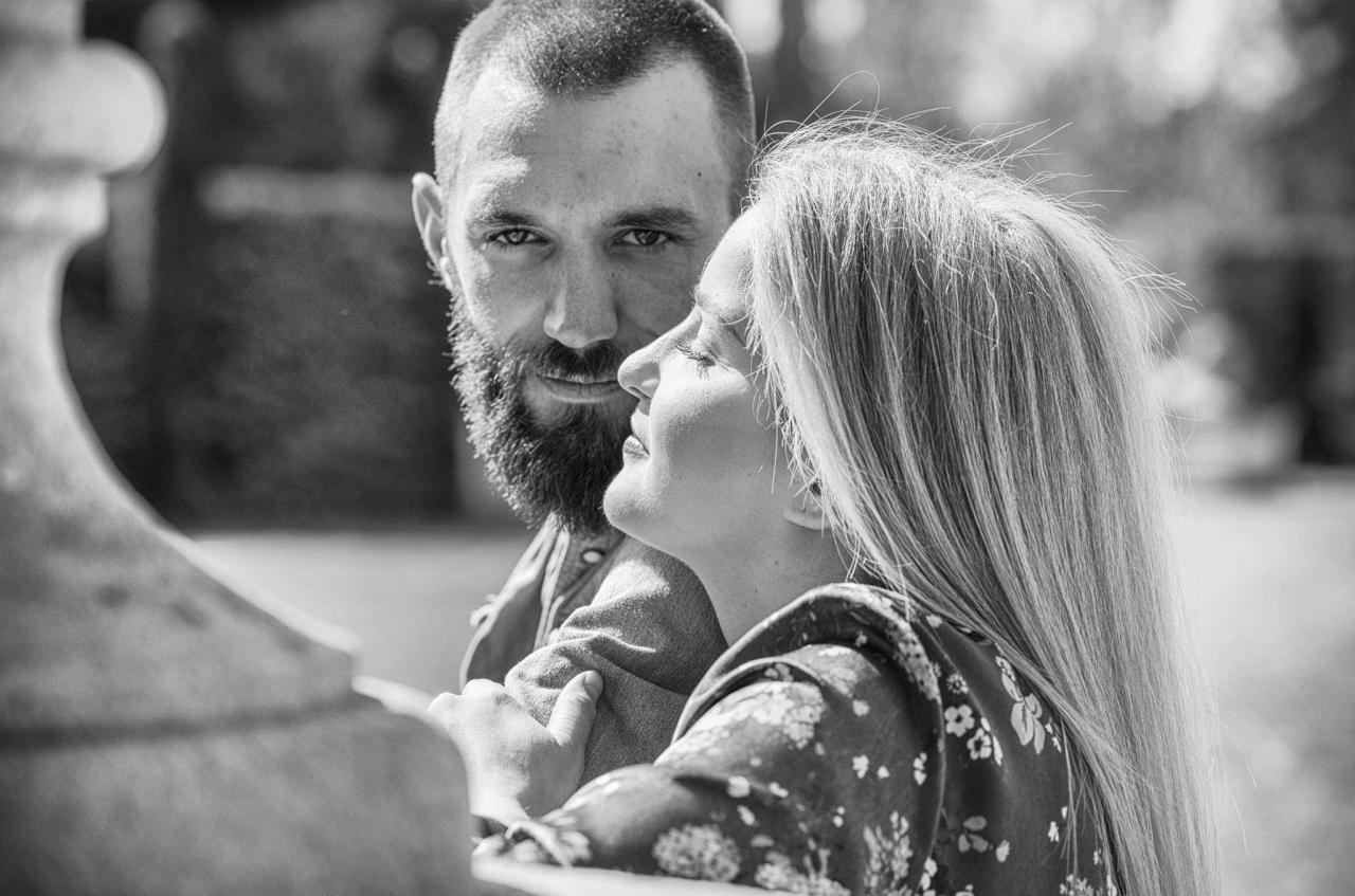Czarno-białe zbliżenie pary: brodaty mężczyzna patrzy w obiektyw, podczas gdy kobieta z długimi włosami spogląda w górę, opierając się o kamienny element architektoniczny.