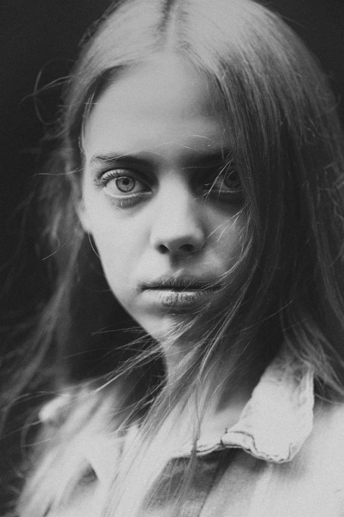 Czarno-biały portret młodej kobiety z długimi, rozwianymi włosami, o poważnym spojrzeniu skierowanym wprost w obiektyw, z widocznym delikatnym światłem padającym na twarz.