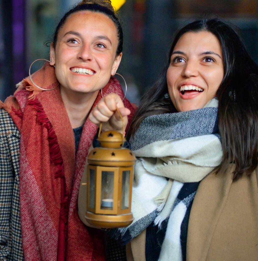 Dwie uśmiechnięte kobiety, jedna trzymająca złotą latarenkę, patrzą w górę, ubrane w szale w kratę i płaszcze.