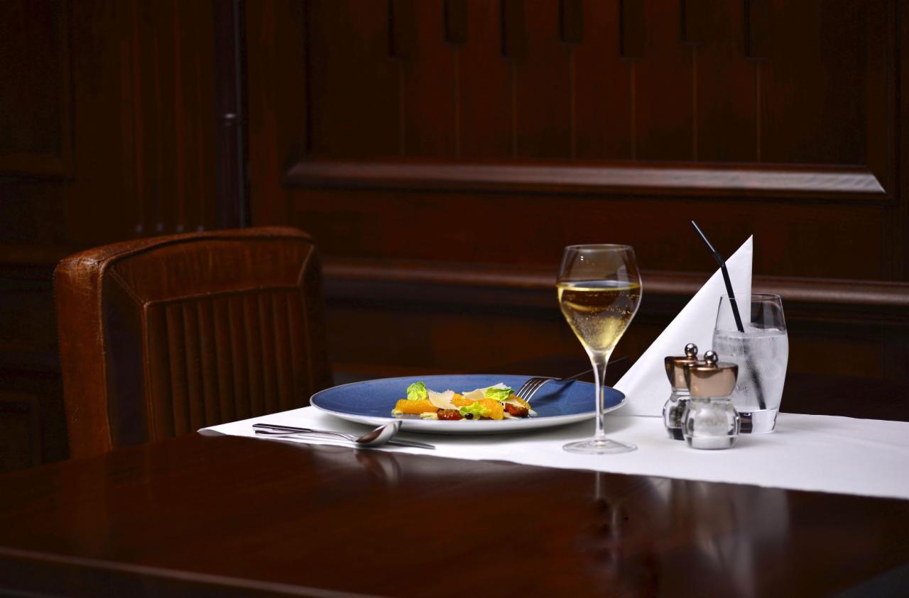 Elegancko nakryty stół w restauracji: niebieski talerz z sałatką, kieliszek szampana, szklanka wody z lodem i słomką, solniczka i pieprzniczka na białym obrusie, ciemne drewniane tło.