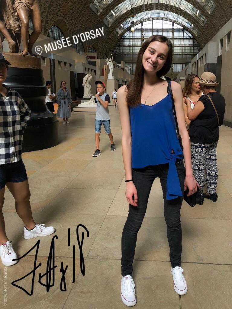 Uśmiechnięta młoda kobieta w niebieskiej bluzce i czarnych dżinsach pozuje na tle wnętrza Musée d'Orsay w Paryżu, z widocznymi rzeźbami i innymi zwiedzającymi.