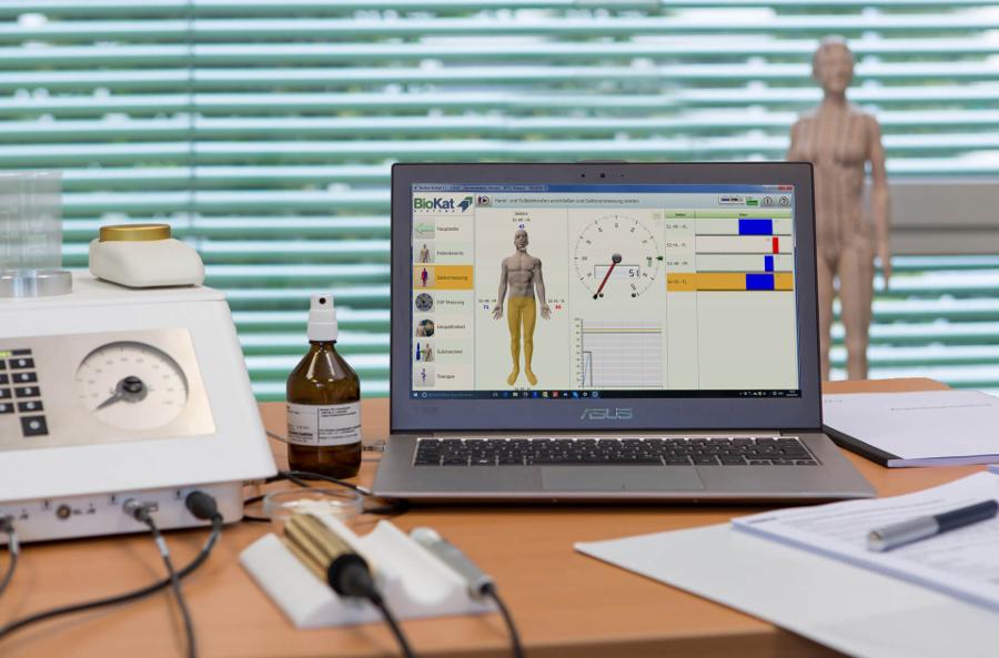 Stanowisko diagnostyczne z laptopem wyświetlającym wizualizację ciała, urządzeniem pomiarowym, butelką z atomizerem i modelem anatomicznym w tle.