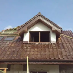 Brązowa dachówka ceramiczna na dachu z oknami dachowymi i lukarną podczas montażu, widoczne deski łat i fragment rusztowania.