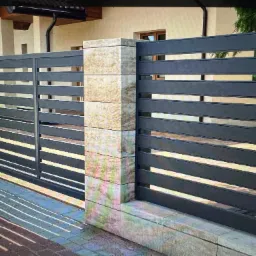 Nowoczesne, poziome przęsła ogrodzeniowe w kolorze grafitowym, osadzone na słupkach z dekoracyjnego kamienia w Warszawie. Widoczne cienie rzucane przez ogrodzenie na chodnik.