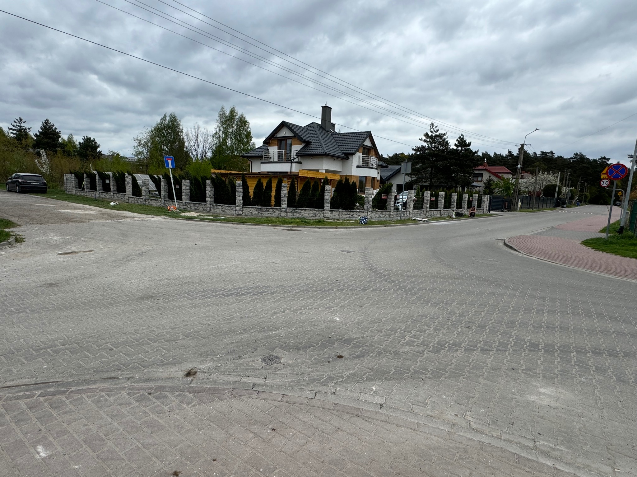Kamienny płot z tujami otaczający posesję z nowoczesnym domem jednorodzinnym, widok z ulicy w pochmurny dzień.