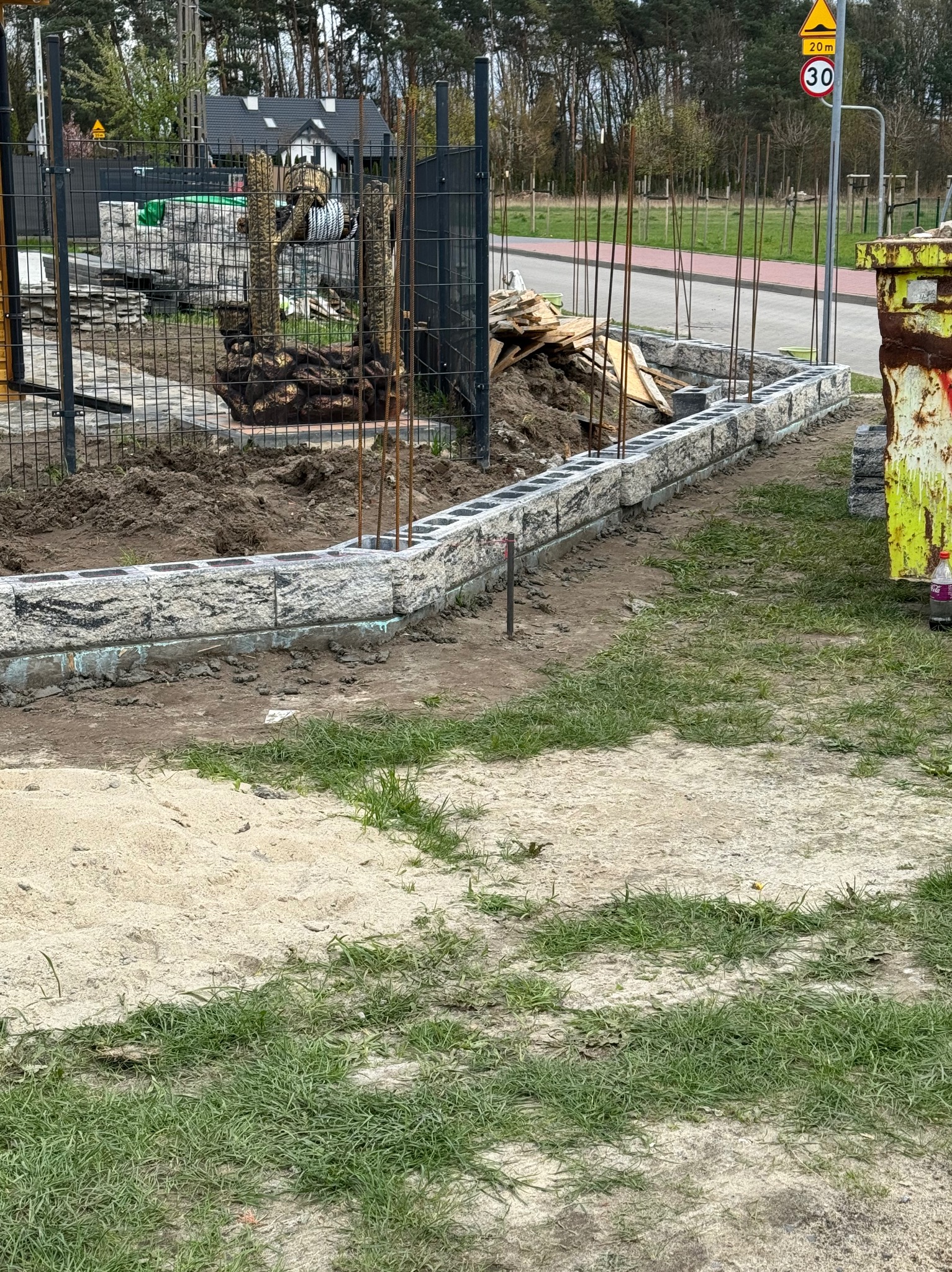 Wznoszenie murku oporowego z bloków betonowych o fakturze kamienia, z wystającymi prętami zbrojeniowymi, widoczne przygotowane podłoże i ogrodzenie panelowe w tle.