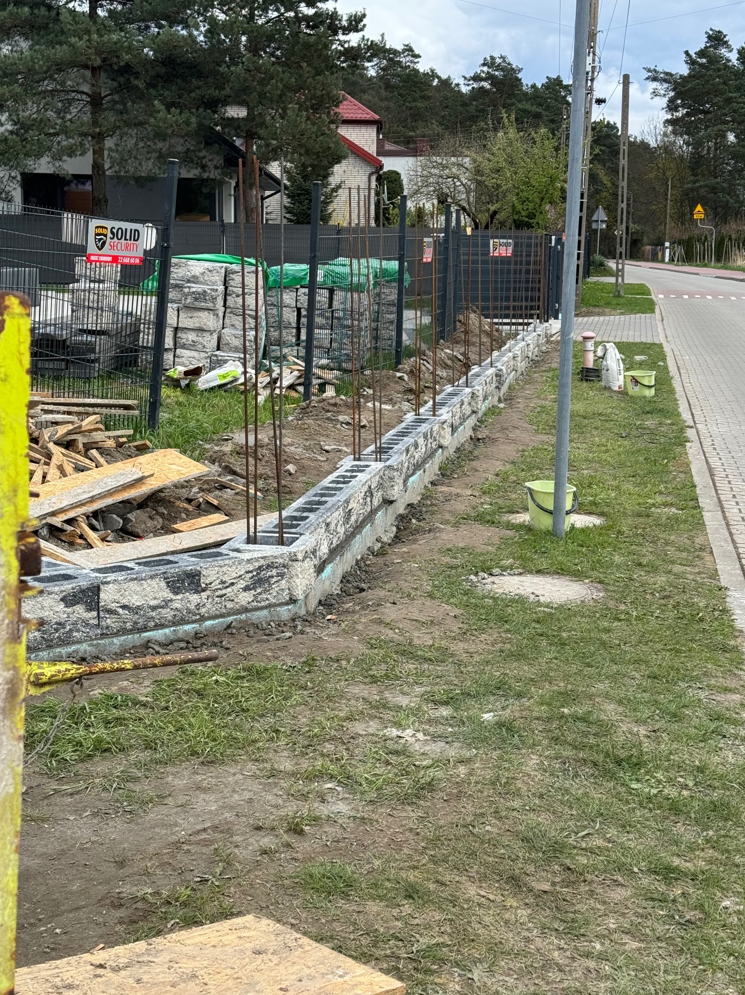 Budowa podmurówki ogrodzenia z szarego bloczka betonowego, wystające pręty zbrojeniowe, widoczne materiały budowlane i narzędzia w tle, teren budowy.